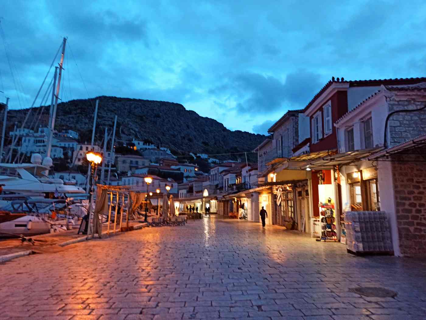 Hydra's waterfront at dusk, the lamplit lanes and harbour bars coming to life