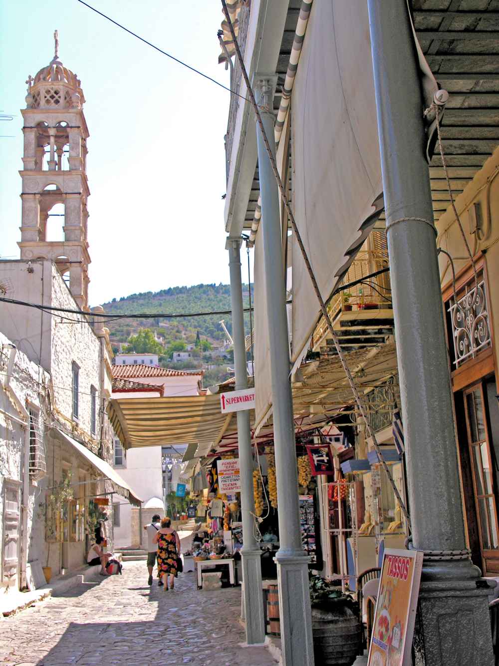 Hydra shopping: a lane of shops under awnings in the town, with the bell tower beyond