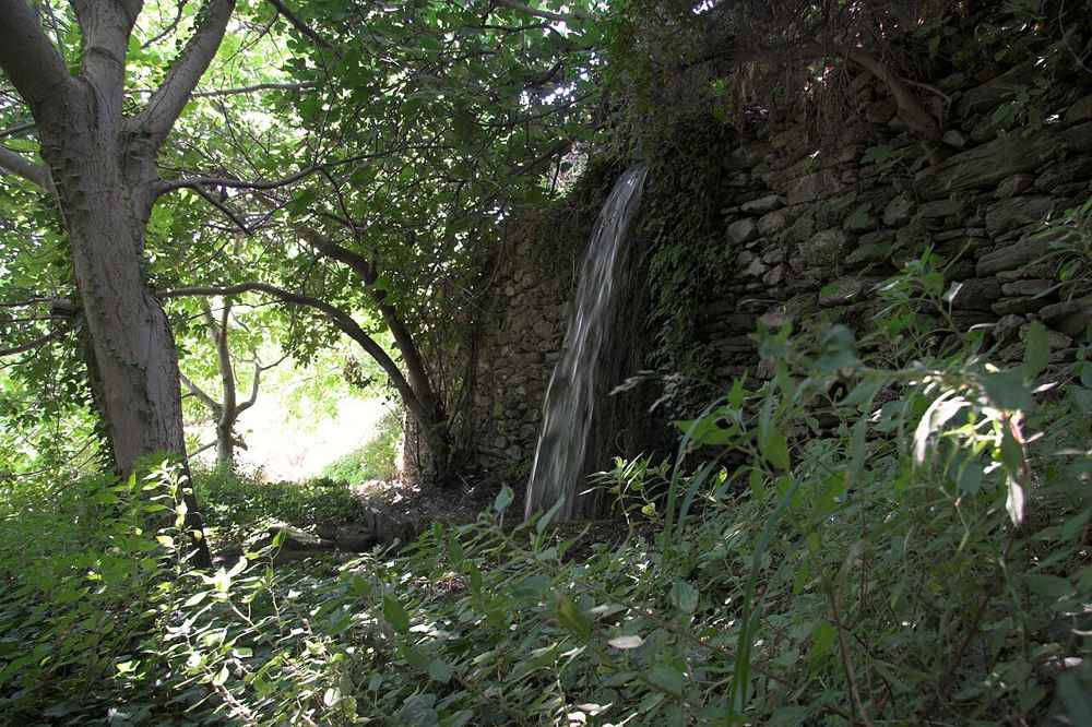 A cascade tumbling past an old stone wall amid the green plane trees of the Andros interior