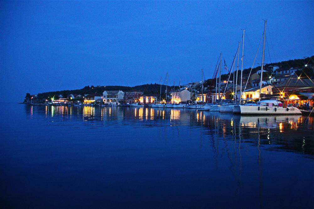 Sailing yachts moored along the lit waterfront of Gaios harbour on Paxos in the evening