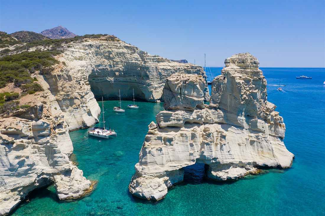 Sailing yachts and a catamaran anchored in the turquoise water below the white sea caves of Kleftiko on a Milos yacht charter
