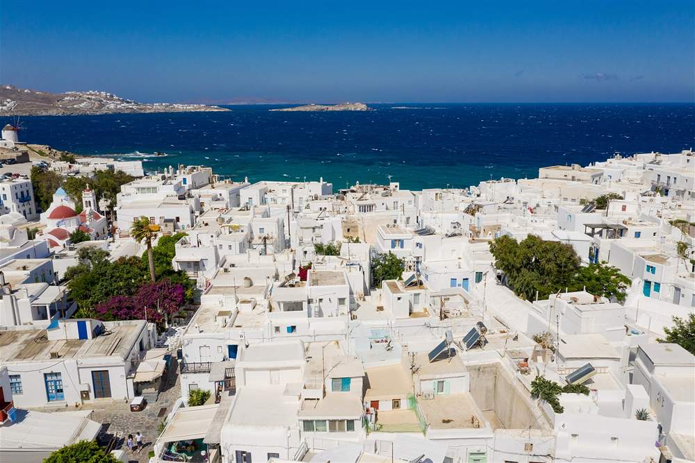 The whitewashed rooftops of Mykonos Town from above with a windmill and the Aegean beyond, a stop on the flight route