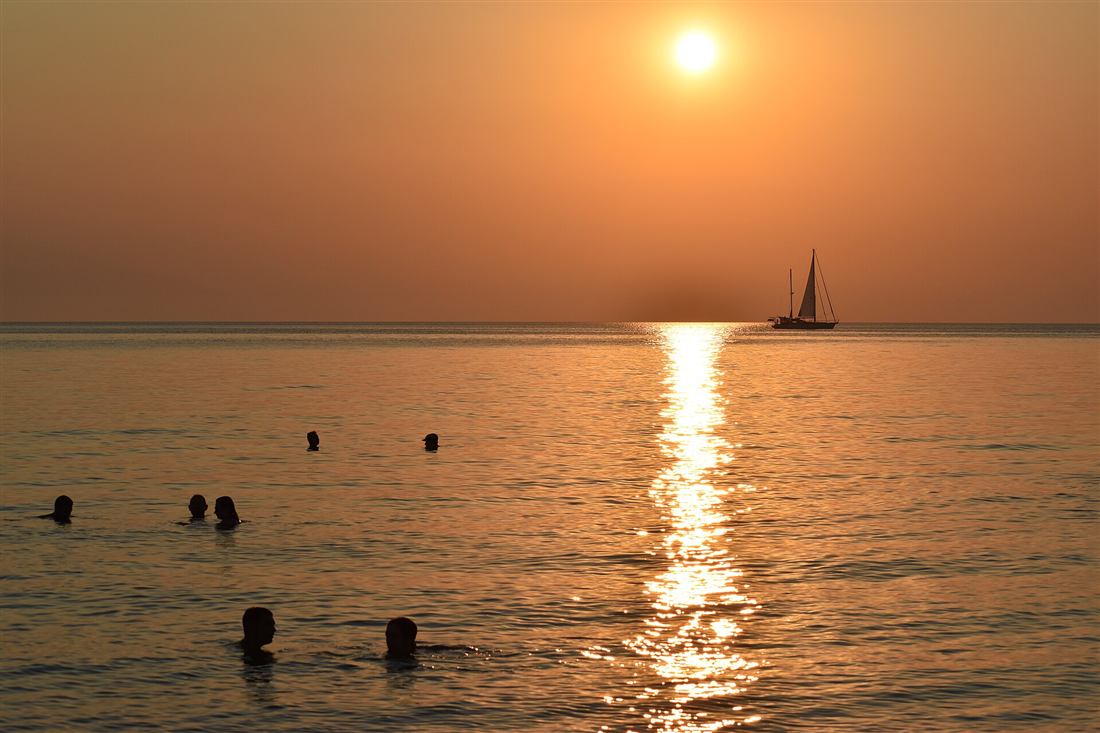 A sailing yacht on the horizon and swimmers in the water as the sun drops over the Aegean