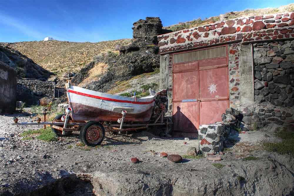 A small wooden boat on its trailer beside a volcanic-stone boathouse on the Santorini coast