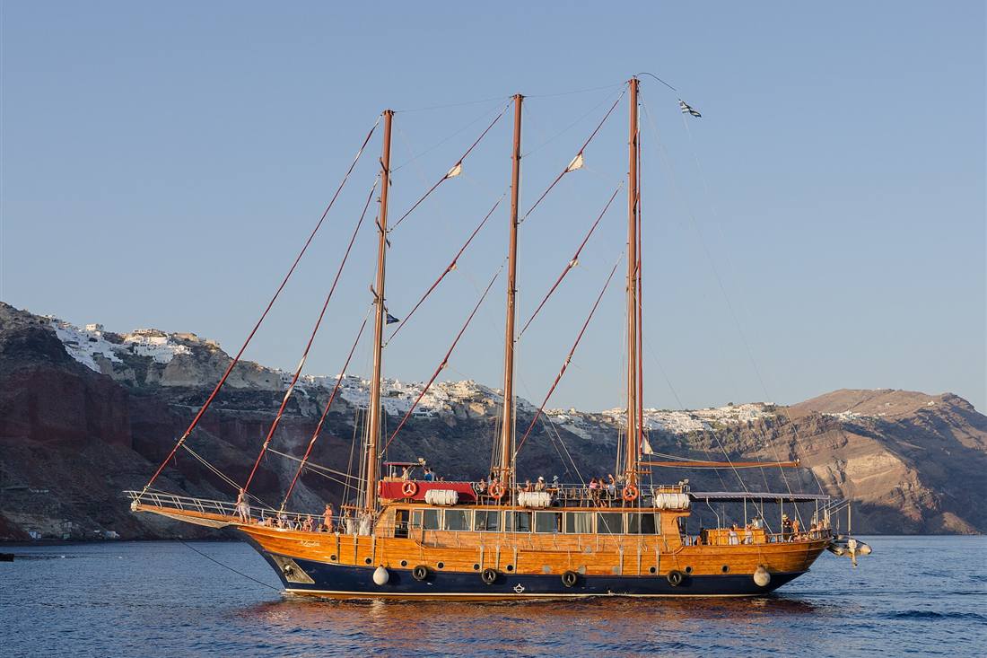 A wooden three-masted tour boat crossing the Santorini caldera below Oia in the evening light, the vessel a Santorini sunset cruise sails on