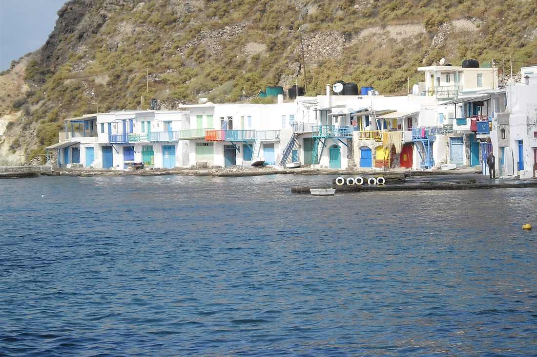 Whitewashed houses with blue, green, red and yellow doors and balconies in a row along a shore, with a dry hillside behind and blue sea in front