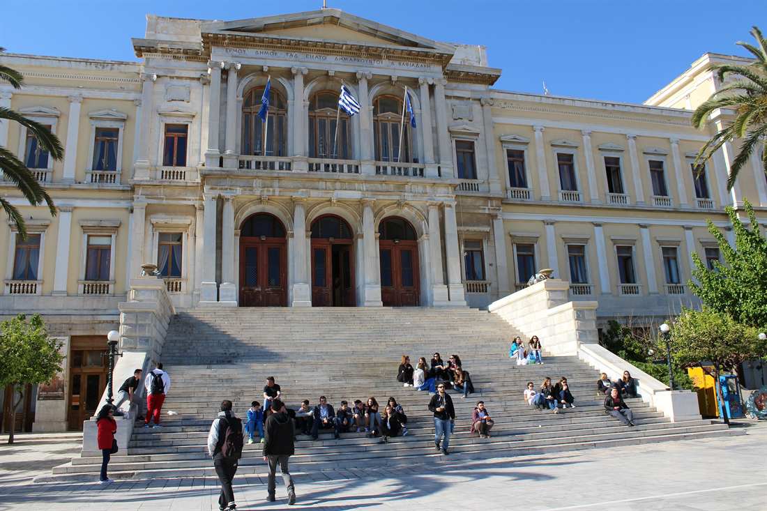 Neoclassical town hall with tall columns and arched doors, Greek and blue EU flags, and a wide marble staircase with people sitting on the steps under a clear blue sky