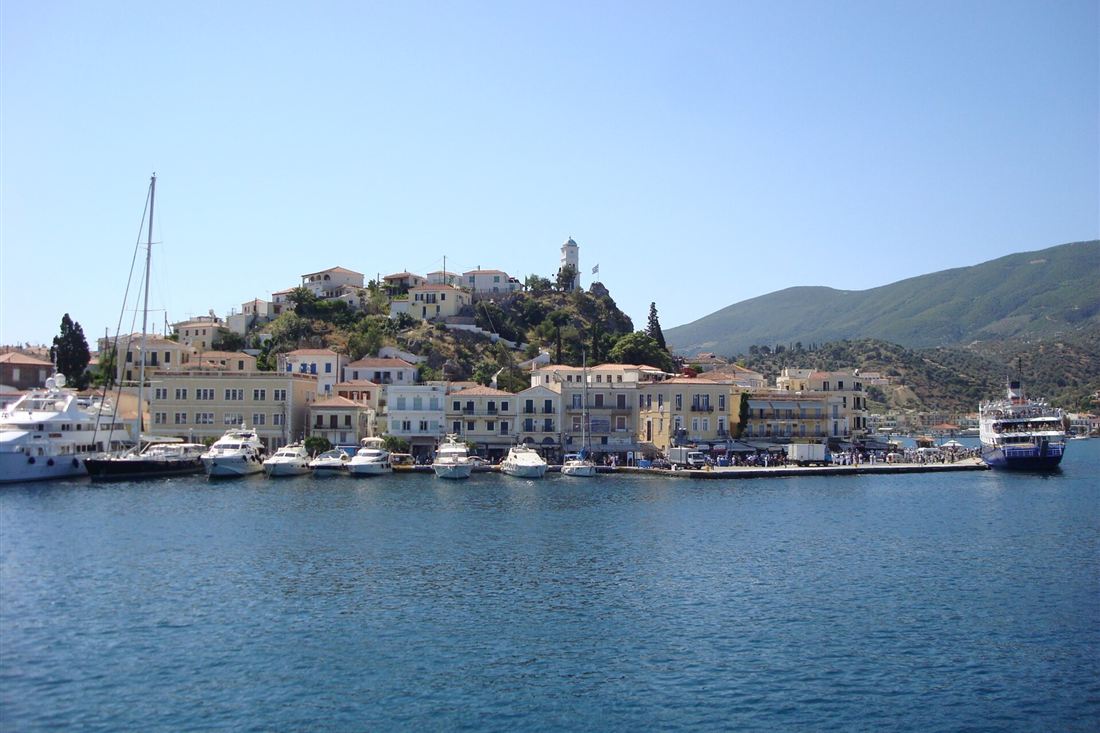 Poros town and its hilltop clock tower seen from the sea, with yachts along the waterfront under a clear sky.