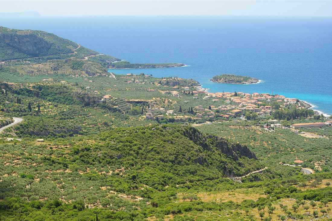 The coast of Kardamyli in the Mani, with olive groves, red-roofed houses and a small islet in a blue sea.