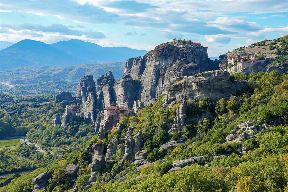 Four Meteora monasteries, St Nicholas Anapafsas, Roussanou, Great Meteoron and Varlaam, on their rock pillars above green slopes with mountains behind.