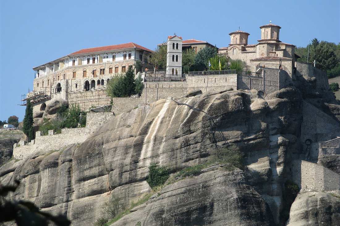 The Varlaam monastery with its domed church and bell tower on top of a sheer grey rock at Meteora under a clear blue sky.