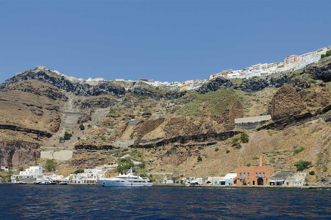 The layered caldera cliffs below Fira on Santorini, with the old port at the waterline and the white town along the rim under a clear sky.