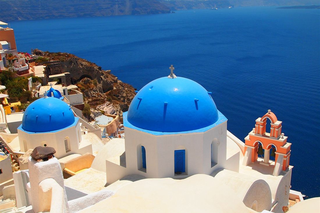 The blue domes and the pink bell tower of an Oia church on Santorini above the deep blue caldera on a clear day.