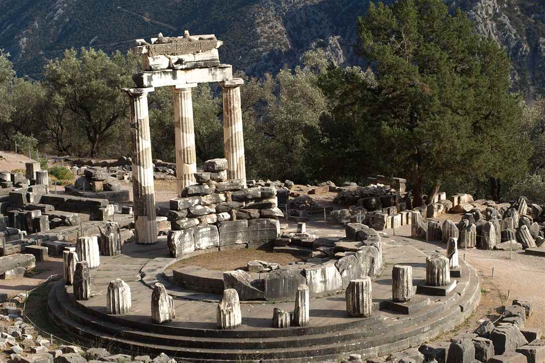 The round Tholos of Athena Pronaia at Delphi, with three restored columns on its stepped platform and the wooded slopes of Mount Parnassus behind.