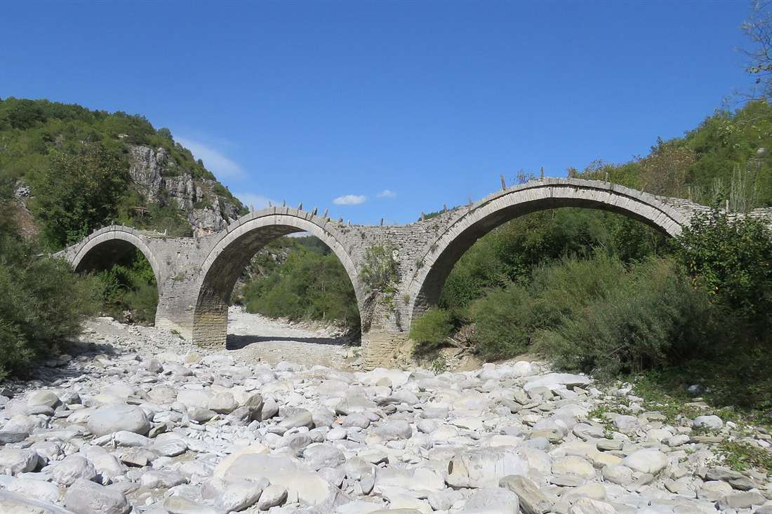 The three-arched Kalogeriko stone bridge in Zagori over a dry pebbled riverbed under a deep blue sky.