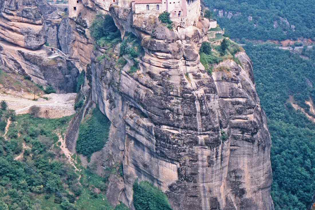 The Great Meteoron monastery on top of its sheer sandstone pillar at Meteora, seen from above with green slopes below.