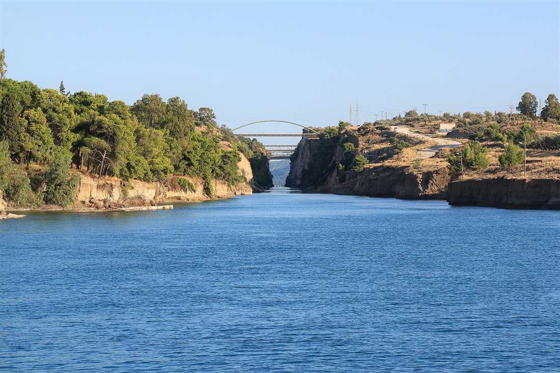 The Corinth Canal opening into the sea between wooded banks, with its road bridges spanning the cut under a clear sky.