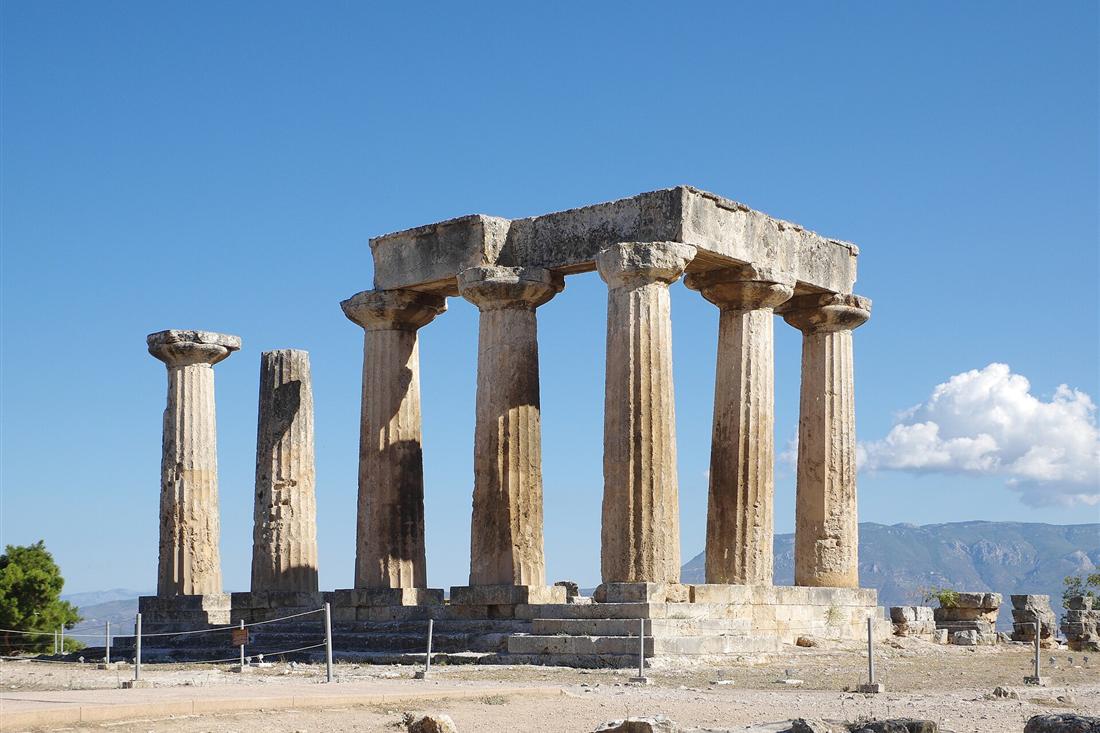 The Doric columns of the Temple of Apollo at Ancient Corinth under a clear blue sky, with mountains on the horizon.