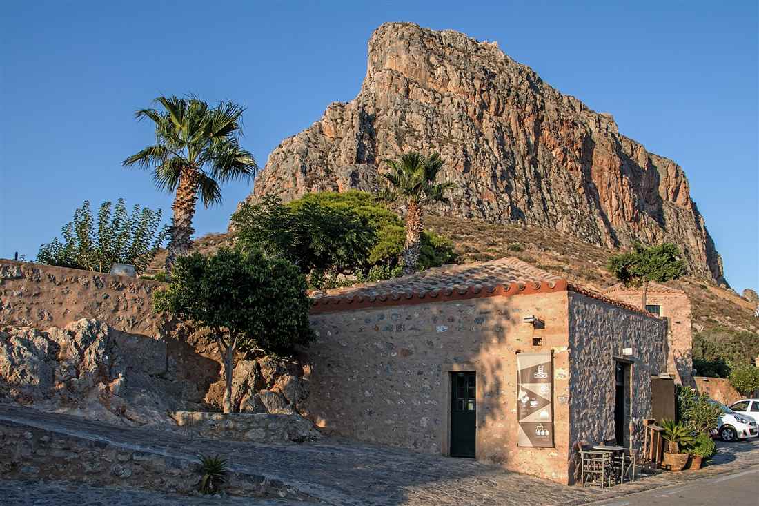 The rock of Monemvasia rising above a stone house with a tiled roof and palm trees, under a clear blue sky.