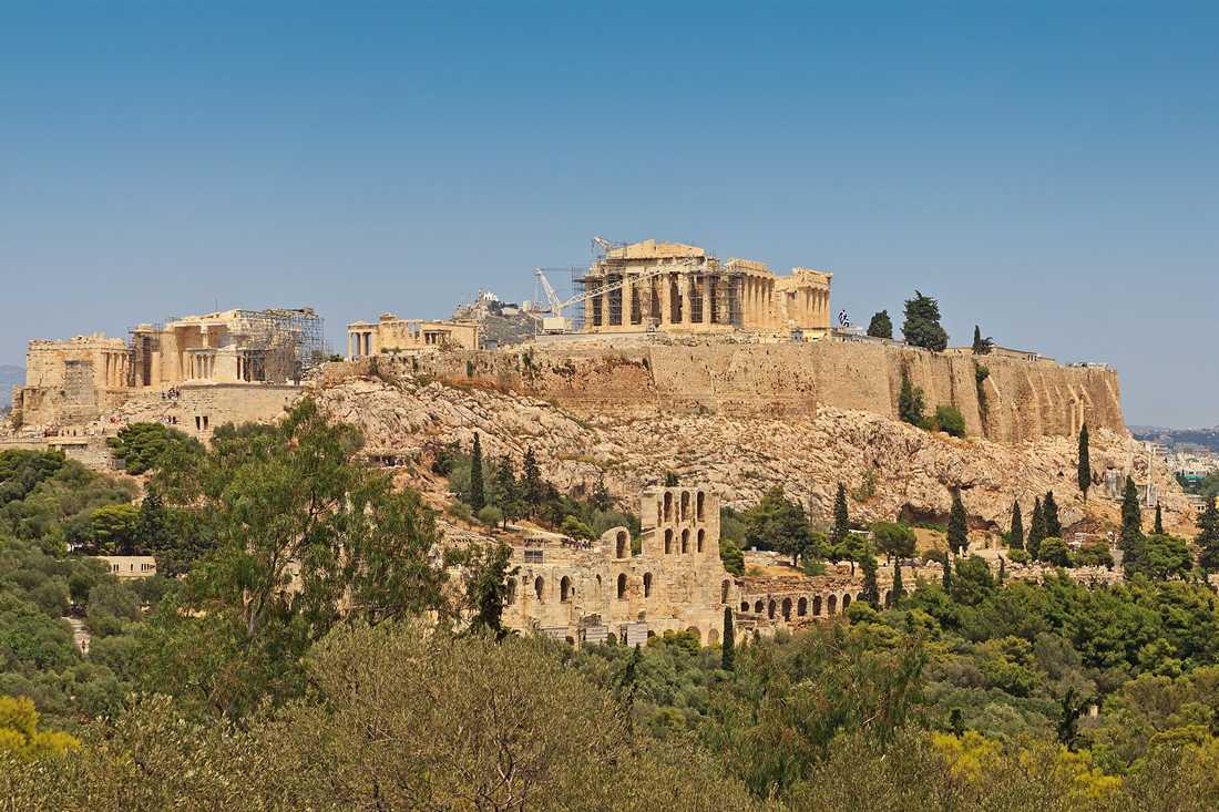The Acropolis of Athens on its rock above green slopes, with the Parthenon at the top and the Odeon of Herodes Atticus below, under a clear blue sky.