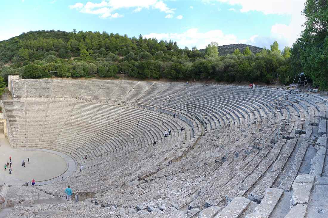 The ancient theatre of Epidaurus with its curved limestone tiers set into a green wooded hillside under a blue sky.