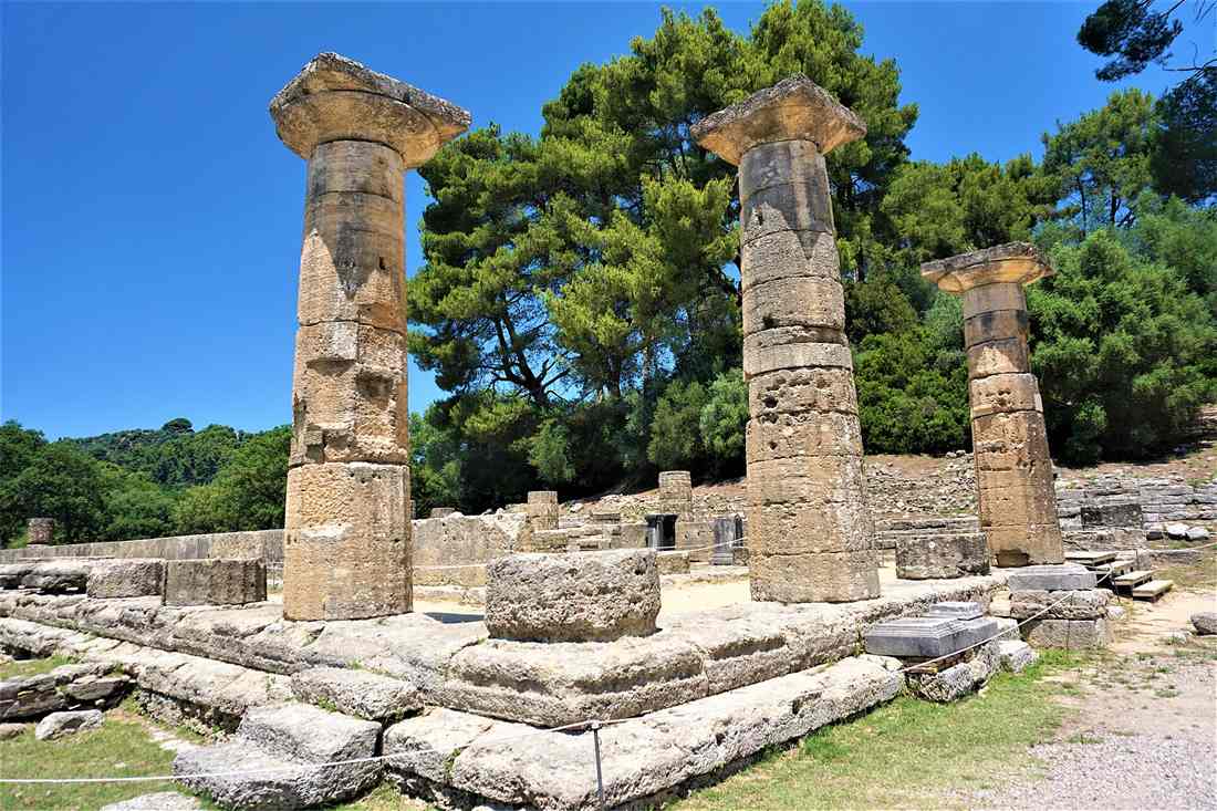 The Doric columns of the Temple of Hera at Ancient Olympia standing on their stone platform under a clear blue sky, with pine trees behind.