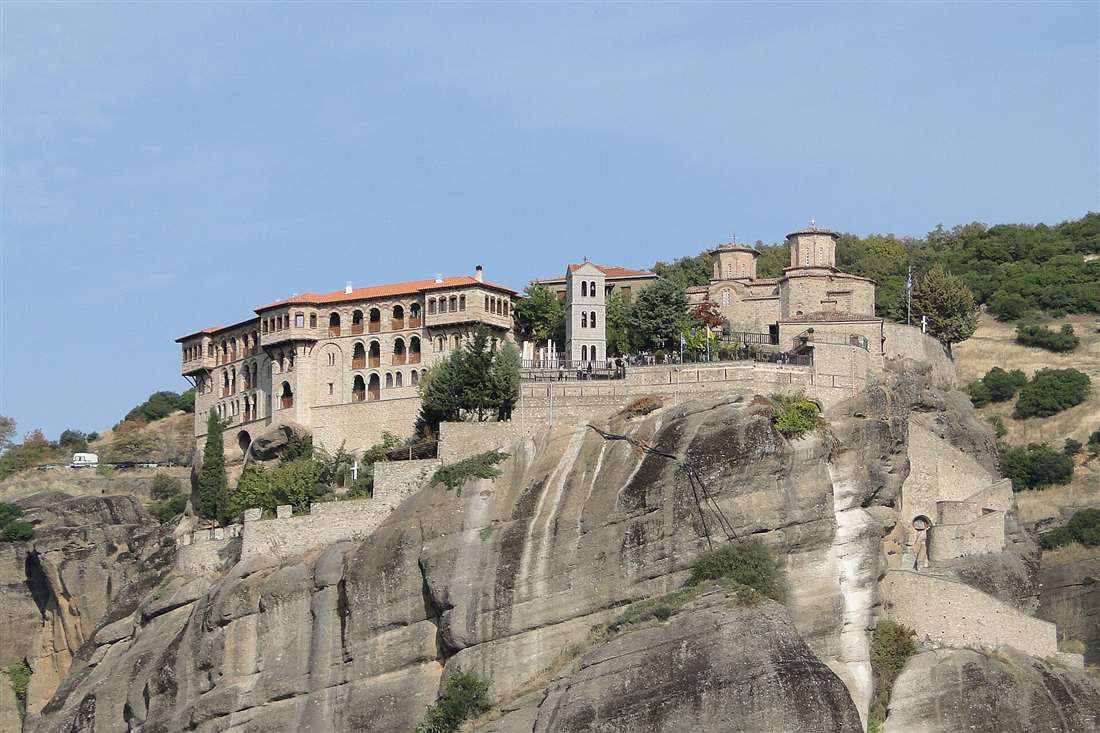 The Great Meteoron Monastery on its sandstone rock at Meteora, with stone buildings, a bell tower and a domed church under a clear blue sky.