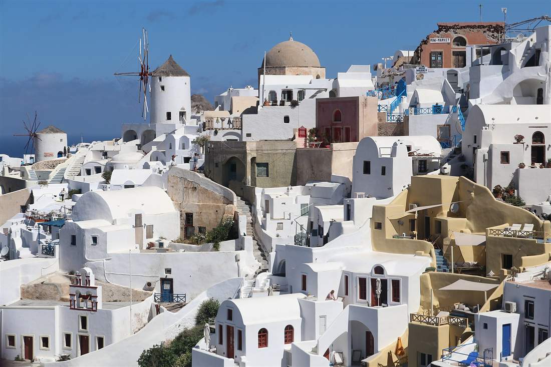 White houses, a domed church and windmills of Oia on the Santorini cliffs under a clear blue sky.