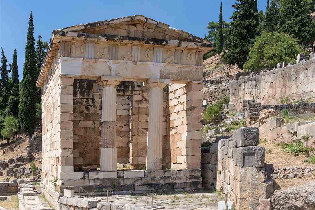 The marble Athenian Treasury at Delphi, with Doric columns, carved metopes and cypress trees under a blue sky.
