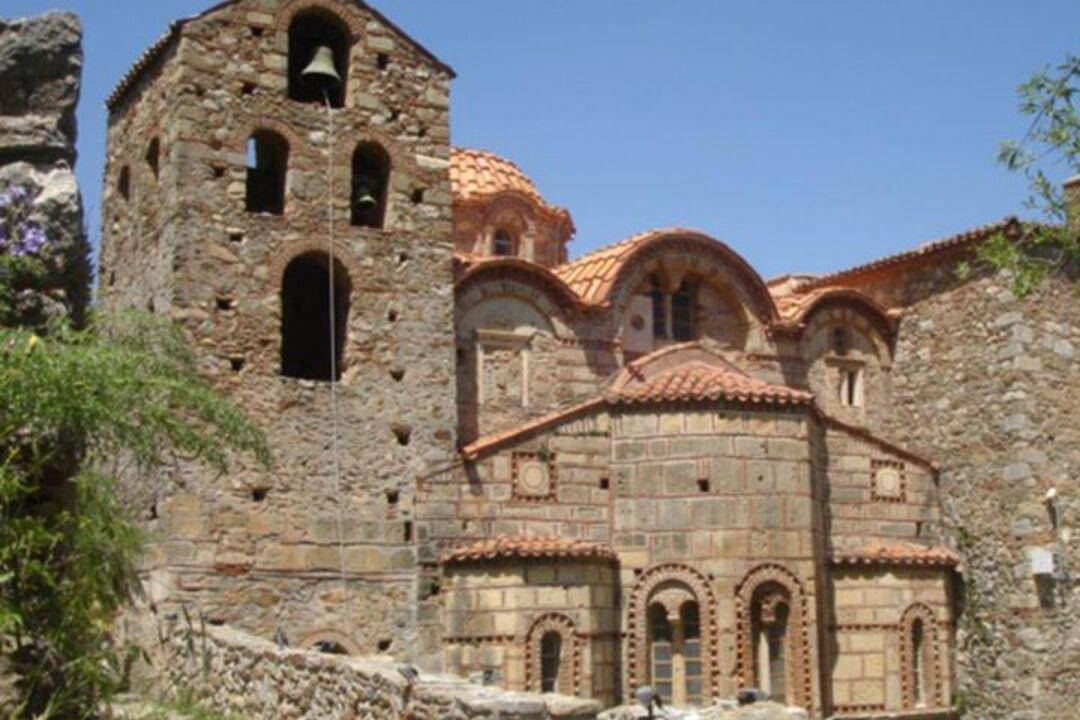 A Byzantine church with a stone bell tower and red-tiled domes at Mystras, under a clear blue sky.