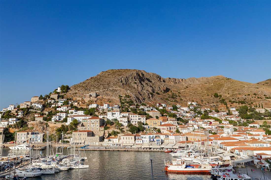 Hydra harbour with yachts and boats moored below stone houses climbing the rocky hill, under a clear blue sky.