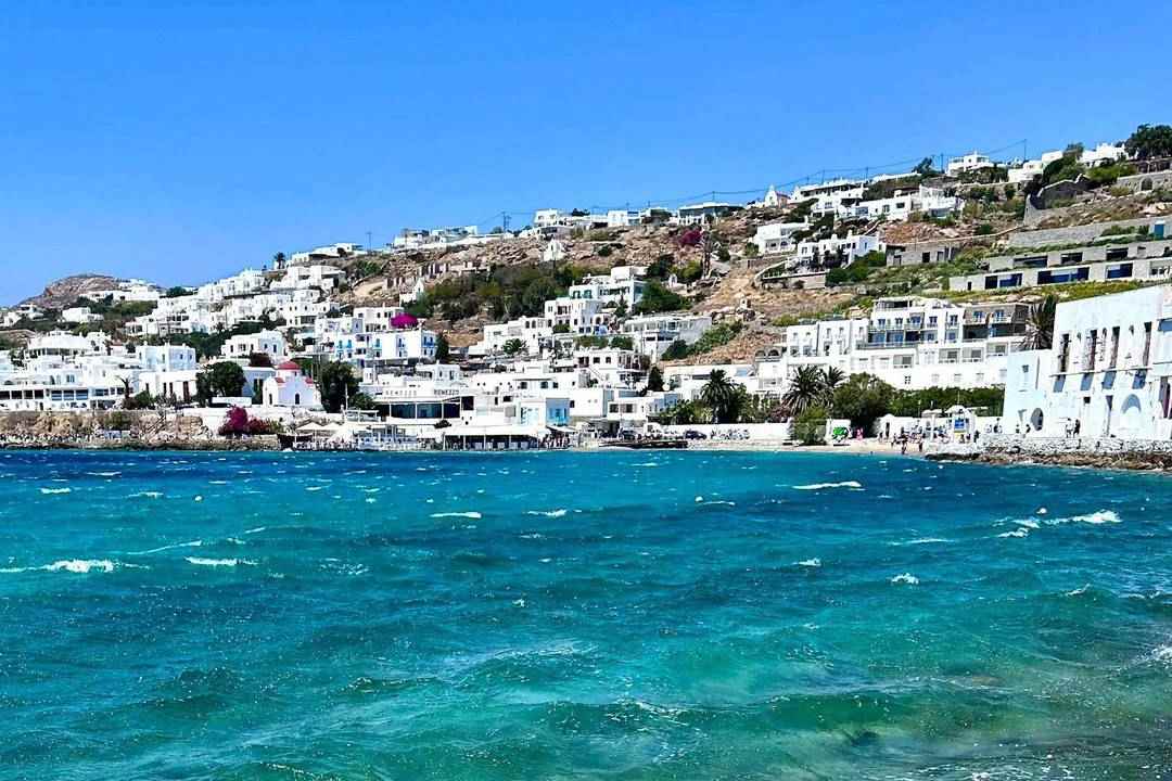 White houses of Mykonos town along the waterfront, with turquoise sea in the foreground.
