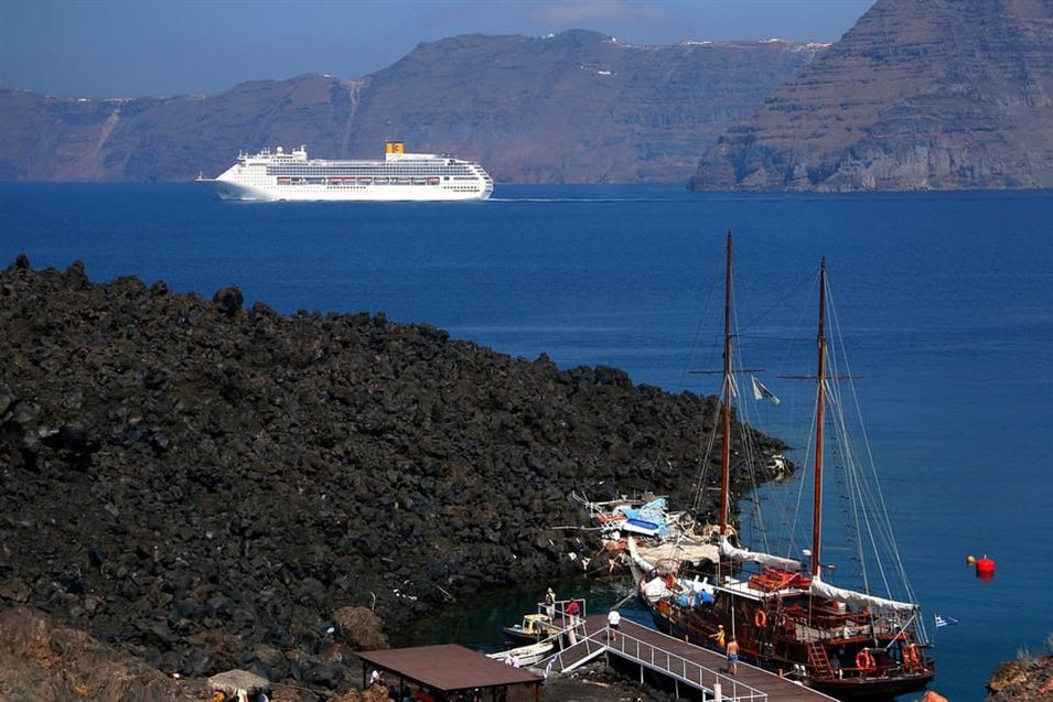Traditional wooden sailing boats moored at the dark lava shore of Nea Kameni, with the Santorini caldera cliffs behind.