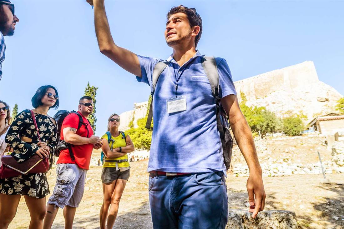 A tour guide in a blue polo shirt with a lanyard badge raises one arm to point while a group of visitors in sunglasses stands around him, with a pale stone hill wall and green trees behind under a clear blue sky