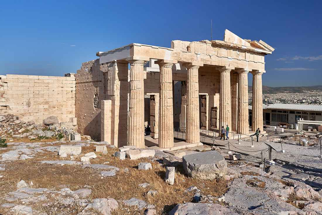 The Propylaea gateway on the Acropolis of Athens, with tall Doric columns, a broken pediment above and a stone wall on the left, and the city of Athens beyond under a deep blue sky