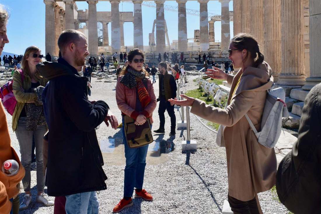 A guide speaking with a small group of visitors on the Acropolis of Athens, with the columns of the Parthenon rising behind them in bright sun under a blue sky