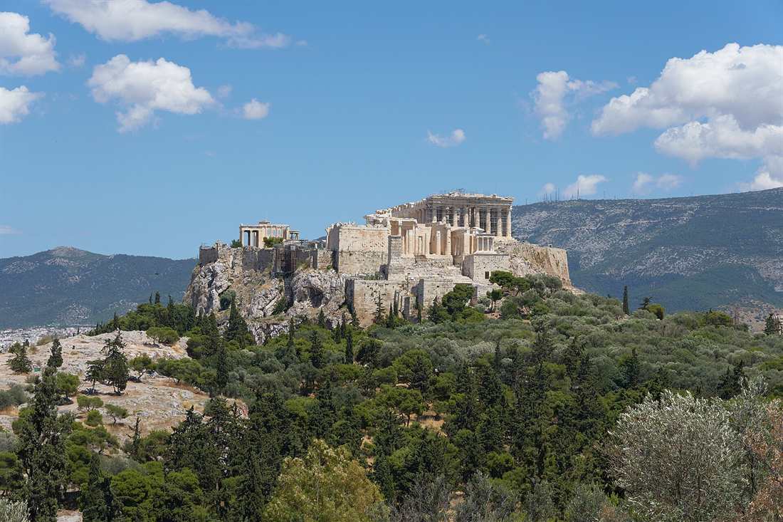 The Acropolis of Athens on its rock with the Parthenon and the Erechtheion on top, above a wooded hill under a blue sky with white clouds