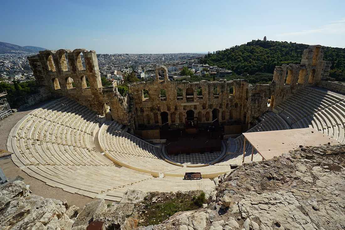 Odeon of Herodes Atticus seen from the Acropolis rock, with the curved stone seating, the arched stage wall and a wooded hill beyond under a clear sky