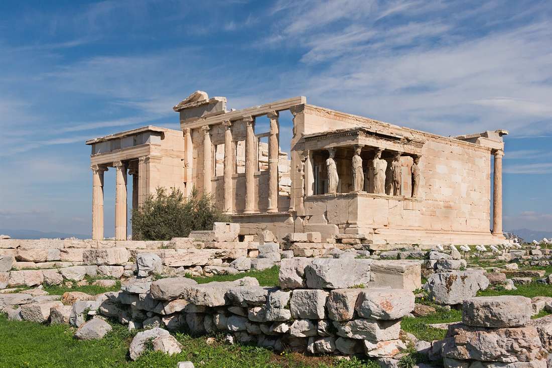 The Erechtheion on the Acropolis of Athens, with the Caryatid Porch on the right and tall Ionic columns in the centre, above marble blocks and grass under a blue sky with thin clouds