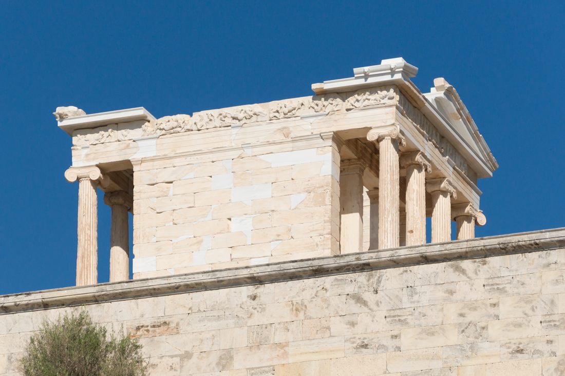 The Temple of Athena Nike on its bastion on the Acropolis of Athens, with Ionic columns and a carved frieze above the bastion wall under a clear deep blue sky