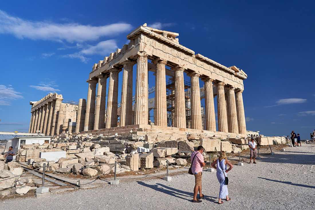 The east front of the Parthenon on the Acropolis of Athens, a row of tall Doric columns under a stone pediment against a deep blue sky with white clouds, with two visitors walking past roped-off marble blocks in the foreground