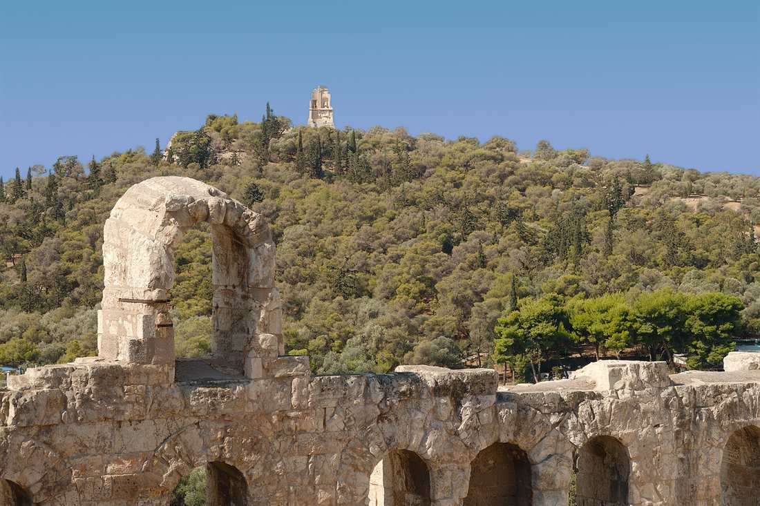 A weathered stone arch and arcade wall in the foreground, with a wooded hill rising behind it and a small stone monument on the summit under a clear blue sky
