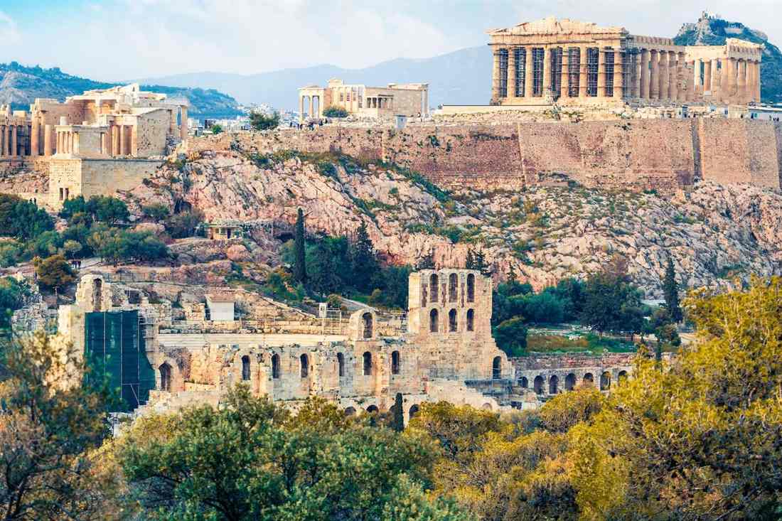 The Acropolis of Athens seen from a wooded hill, with the Propylaea gateway on the left, the Erechtheion in the centre, the Parthenon on the right above the rock wall, and the arched stone facade of the Odeon of Herodes Atticus below