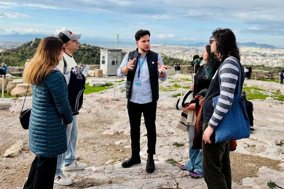 A guide in a black vest with a blue lanyard badge speaks with a group of visitors on the pale rock of the Acropolis, with a green hillside and the city of Athens behind under a blue sky with thin clouds