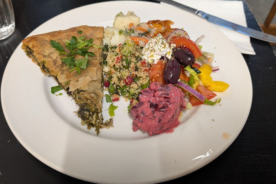 A slice of baked Greek spinach pie topped with fresh parsley on a white plate beside salad with olives and feta