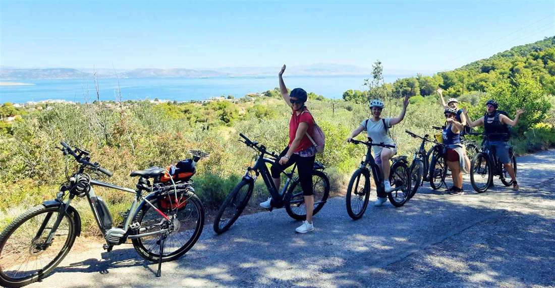 A group of riders with e-bikes waving on a country road on Aegina, with the Saronic Gulf behind them