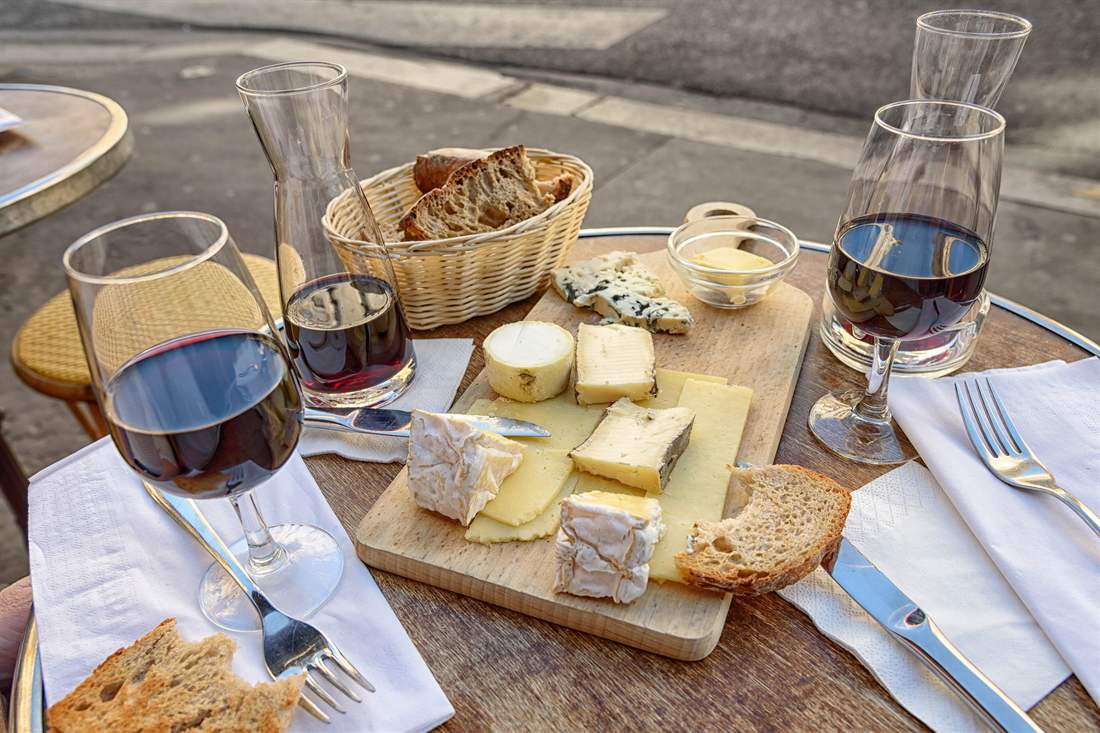 Glass of red wine beside a wooden board of sliced cheeses and bread with carafes of red wine on a cafe table