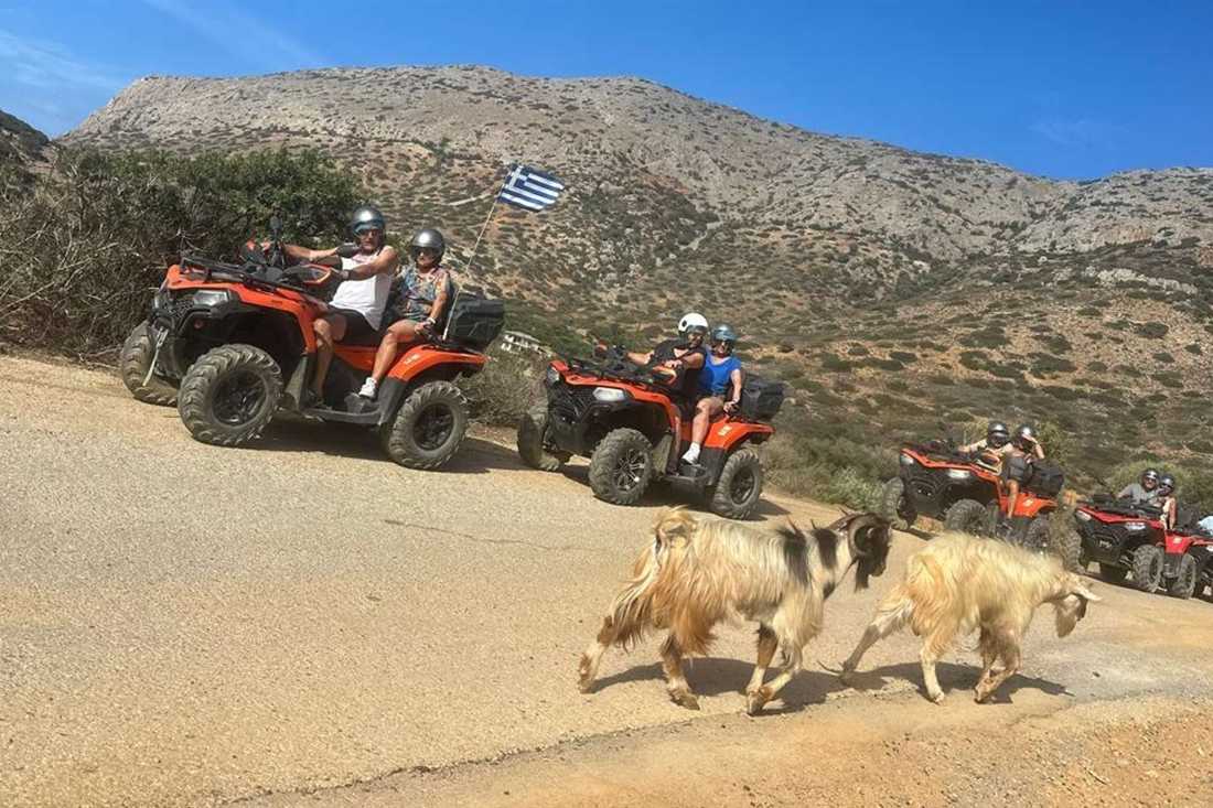 Convoy of orange quad bikes with a Greek flag on a dirt mountain road in Crete, two goats walking past in front