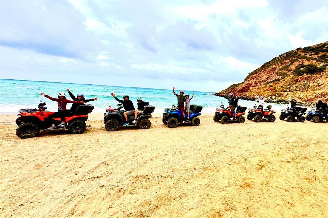 Row of quad bikes with riders waving on a sandy beach beside a turquoise sea and a red-brown hillside
