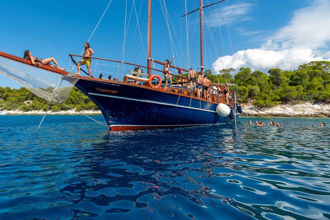 Blue wooden sailing gulet anchored in clear blue water beside a pine-covered rocky shore, with passengers on deck and swimmers in the water